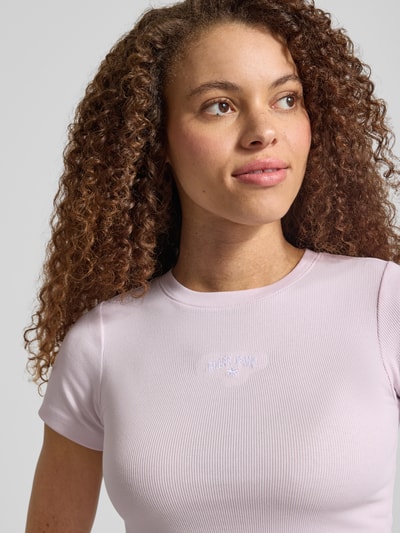Frauenmodel trägt T-Shirt in Ripp-Optik mit Logo von Guess Jeans in Hellrosa, Slim Fit, Baumwoll-Elasthan-Mix, Nahaufnahme Oberkörper.