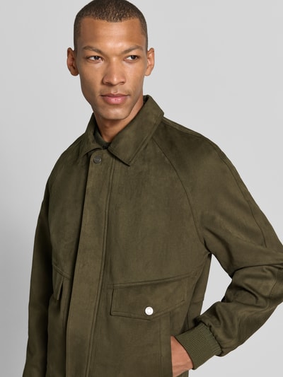 Męski model nosi kurtkę w imitacji zamszu Model 'AVIATOR' od GUESS, elastyczna mieszanka materiałów, krój regular fit, wykładany kołnierz, plisa z zatrzaskami, kieszenie z patką, elastyczne wykończenia, kolor ciemnozielony, gładka tekstura, półzbliżenie, widok z przodu.