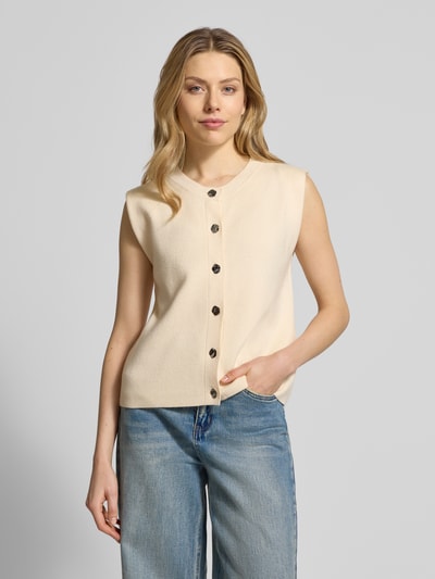 Frauenmodel trägt Regular Fit Strickjacke aus Viskose-Mix Modell 'GOLD' von Vero Moda in Beige, einreihige Knopfleiste, unifarben, Strickoptik, ohne Ärmel, gerippter Abschluss, Nahaufnahme Oberkörper.