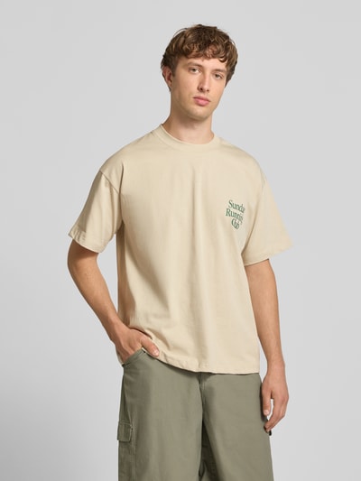 Męski model nosi T-Shirt mit Print und Rundhalsausschnitt od ANOTHER COTTON LAB z bawełny, krój relaxed fit, nadruk, okrągły dekolt, długość tyłu w rozmiarze M 69 cm, beżowy kolor, prosta faktura, całkowity widok z przodu.