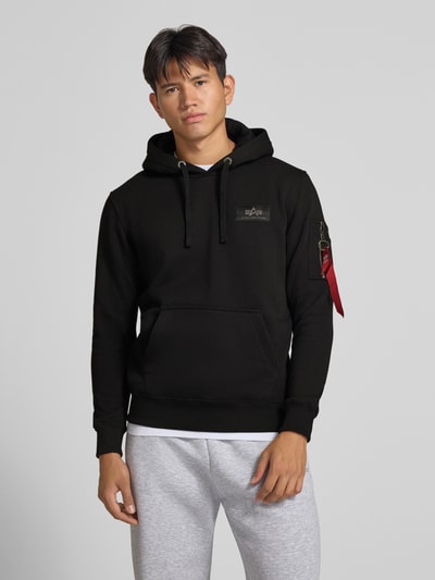 Männermodel trägt schwarzen Hoodie mit Känguru-Tasche und rückseitigem Label-Print von Alpha Industries, Baumwoll-Mix, Regular Fit, Ganzkörperansicht von vorne.