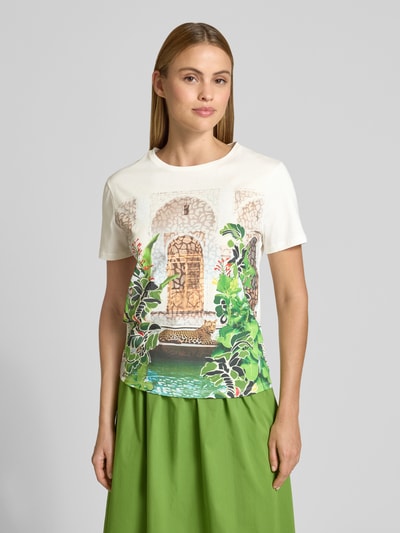Kobiece model prezentuje Regular Fit T-Shirt mit Allover-Motiv-Print von Marc Cain z bawełny, okrągły dekolt, długość tyłu 56 cm, przód w jasnych kolorach, motyw zielonych liści i motyw architektoniczny, gładka tekstura, Ganzkörperansicht von vorne.