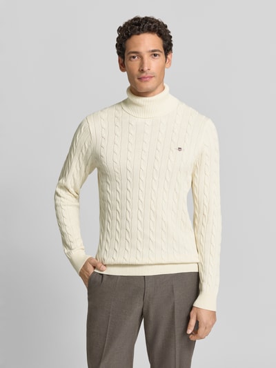 Männermodel trägt Rollkragenpullover mit Logo-Detail von GANT in Creme mit Zopfmuster, gerippten Abschlüssen, reine Baumwolle, Ganzkörperansicht von vorne.