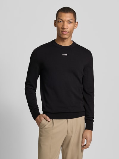 Męski model nosi Regular Fit Strickpullover aus reine Baumwolle Modell 'SAN CASSIUS-C2' od HUGO, czarny, gładka dzianina, crew neck, long sleeve, nadruk z logo, wykończenia ściągaczem, krój regular fit, widok całej sylwetki z przodu.