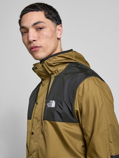 Männermodel trägt Regular Fit Jacke mit logoprint model 'Mountain' van The North Face in olijf en zwart, capuchon met tunnelkoord, steekzakken met ritssluiting, elastische boorden, ruglengte maat M 72 cm, schouderbreedte 48 cm, borstwijdte 53 cm, mouwlengte 70 cm, detailopname bovenlichaam schuin vooraan.