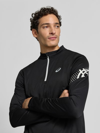 Męski model nosi bluzkę z długim rękawem Langarmshirt mit Troyer-Kragen Modell 'ICON' von ASICS TIGER, czarny z nadrukiem z logo, prosty krój, kołnierz zapinany, Długość tyłu w rozmiarze M: 72 cm, ganzkörperansicht von vorne