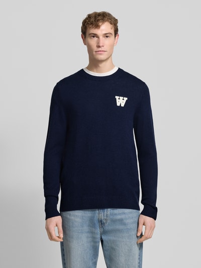 Männermodel trägt Strickpullover aus Lammwolle Modell 'Tay' von Wood Wood in Dunkelblau mit Label-Stitching, Regular-Fit, Rundhalsausschnitt, Ganzkörperansicht von vorne.