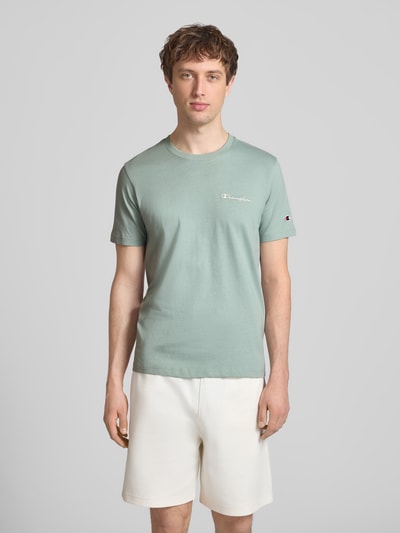 Männermodel trägt T-Shirt mit Label-Stitchings und Rundhalsausschnitt von CHAMPION in Grün, reine Baumwolle, Regular Fit, Ganzkörperansicht von vorne.