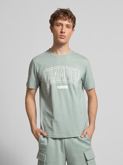 Männermodel trägt T-Shirt mit Logo und Rundhalsausschnitt von CHAMPION in Grün, Reine Baumwolle, Regular-Fit, Label-Stitching, Nahaufnahme Oberkörper.