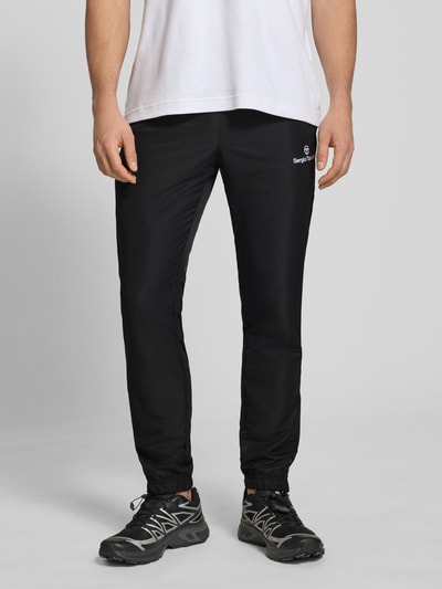 Männermodel trägt schwarze Regular-Fit Tracksuit Hose mit Logo-Stitching von SERGIO TACCHINI, Elastische Abschlüsse, Eingrifftaschen, Nahaufnahme Unterkörper. Modell 'Carson', Bundweite 38 cm, Größe M, glatte Oberfläche.