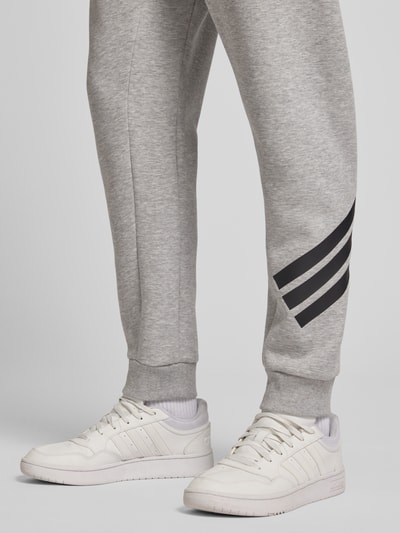 Männermodel trägt Slim Fit Sweathose mit Logo-Print von adidas Sportswear, grauer Material-Mix, elastische Beinabschlüsse, Nahaufnahme Beinbereich.