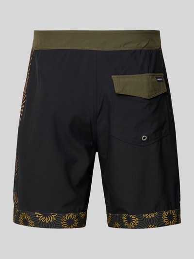 Männermodel trägt Regular Fit Shorts mit Logo-Patch Modell 'Nomad' von Quiksilver mit floralem Print, dunkle Farben, Ganzkörperansicht Rückansicht.