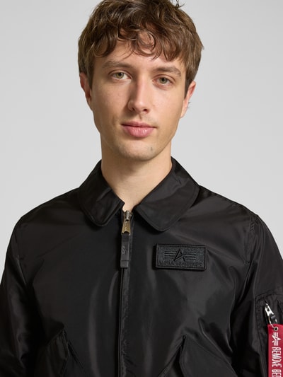 Männermodel trägt schwarze Regular Fit Bomberjacke mit Logo-Patch von Alpha Industries, glatte Textur, Nahaufnahme Oberkörper. Rückenlänge 66 cm, Schulterbreite 44 cm, Brustweite 56 cm, Ärmellänge 66 cm, Umlegekragen, aufgesetzte Taschen, Reißverschluss, elastische Abschlüsse.
