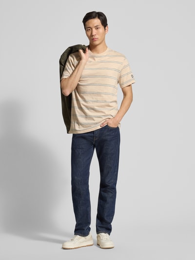 Männermodel trägt T-Shirt mit Rundhalsausschnitt und Label-Patch von Guess, reine Baumwolle, gerade Passform, beige-weiß gestreift, Ganzkörperansicht von vorne.