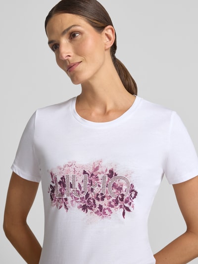 Modelka kobieca nosi Damski t-shirt Regular Fit T-Shirt aus reiner Baumwolle od Liu Jo White, bawełna, krój regular fit, nadruk z logo, obszycie z kamieni stras, okrągły dekolt, długość tyłu w rozmiarze S: 66 cm, widok na górną część ciała z przodu.