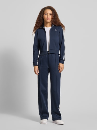 Kobiece model nosi Spodnie typu track pants 'Sportliche Trackhose mit geradem Bein' od REVIEW, granatowe, koronka na bocznym szwie, zaszewka z przodu, wyhaftowane logo na bocznej kieszeni, długość wewnętrzna nogawki w rozmiarze S: 39 cm, widok całej sylwetki z przodu.