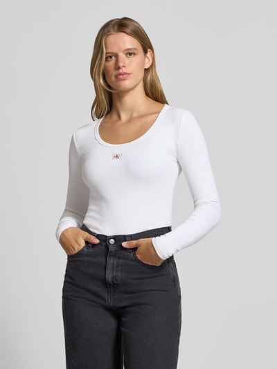 Frauenmodel trägt Slim Fit Langarmshirt aus Baumwoll-Mix von Calvin Klein Jeans in Weiß, glatte Textur, Logo-Applikation aus Metall, Nahaufnahme Oberkörper.