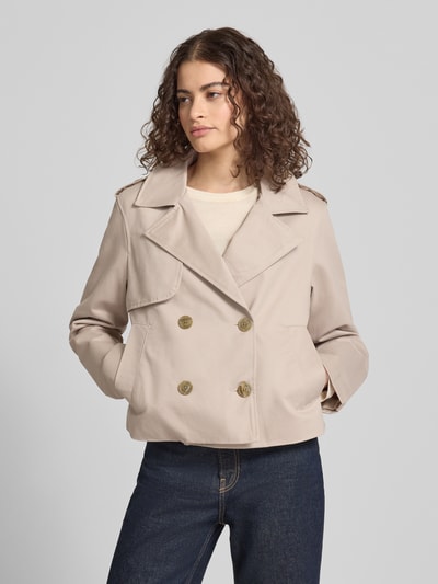 Modelka nosi Regular Fit Trenchcoat mit Umlegekragen od Review, czysta bawełna, krój regular fit, długość tyłu 55 cm, szerokość ramion 45 cm, szerokość w rozmiarze S: 53 cm, długość rękawa 62 cm, jasny beż, kontrastujące guziki, prosty krój, dwurzędowe zapięcie, kieszenie, widok całej sylwetki z przodu.