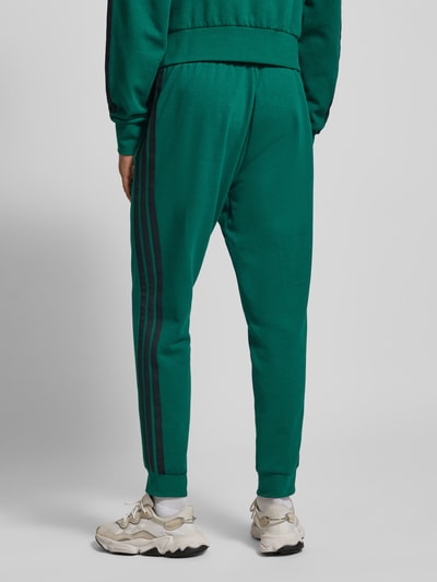 Männermodel trägt Sweatpants mit elastischem Bund von adidas Sportswear aus Baumwoll-Mix mit Viskose-Anteil, Lockerer Schnitt, Tunnelzug, Label-Stitching, grün mit schwarzen Streifen, Innenbeinlänge bei Größe M 76 cm, Ganzkörperansicht Rückansicht.