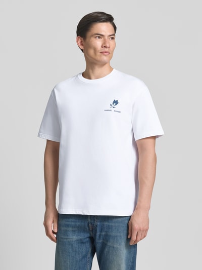 Męski model nosi T-Shirt mit Label-Print Modell 'COHEN' od Samsøe Samsøe z czystej bawełny organicznej, krój relaxed fit, biały, nadruk, okrągły dekolt, długość pleców w rozmiarze M: 71 cm, ujęcie całej sylwetki z przodu.