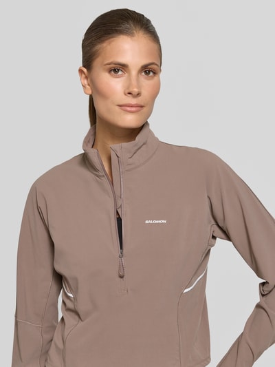 Frauenmodel trägt Cropped Pullover für intensive Anstrengungen und Beschleunigung von Salomon in Taupe mit Logo, Stehkragen, Reißverschluss, Material-Mix mit Stretch-Anteil, ganzkörperansicht von vorne. Rückenlänge bei Größe S: 47 cm.