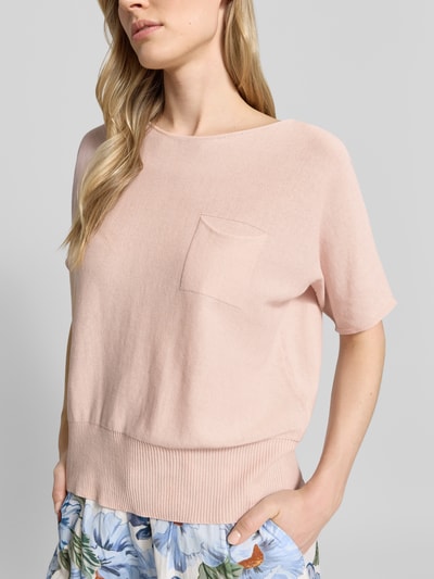 Modelka kobieca ma na sobie Strickpullover mit U-Boot-Ausschnitt und Brusttasche od Oui z czystej bawełny, krój loose fit, jasnoróżowy kolor, półrękawy, tekstura dzianiny, detal przyciętego rąbka oraz kieszonka na piersi, widoczne kwiatowe wzory na spodniach, ujęcie z przodu klatki piersiowej, zbliżenie