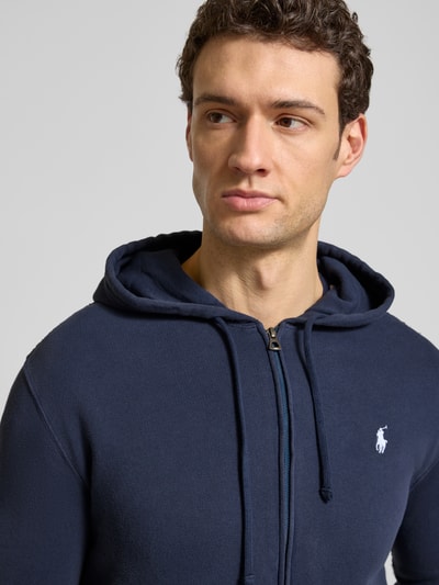 Męski model nosi Regular Fit Sweatjacke aus Baumwoll-Mix von Polo Ralph Lauren, kolor granatowy, logo z przodu, kaptur, materiał bawełniany, bluza rozpinana, krój regular fit, długi rękaw, ściągacze, zbliżenie na górną część ciała