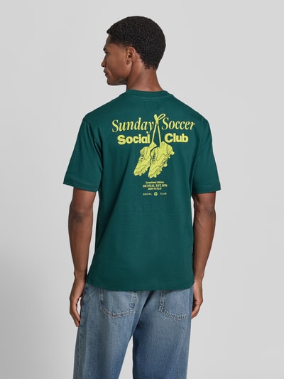 Männermodel trägt T-Shirt mit Statement-Print von MCNEAL in Dunkelgrün mit gelbem Aufdruck, lockerer Schnitt, Ganzkörperansicht Rückansicht, Rückenlänge bei Größe M 66 cm, Rundhalsausschnitt