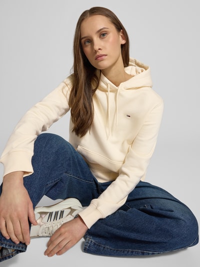 Modelka kobieca ma na sobie Regular Fit Hoodie aus Baumwoll-Mix od Tommy Jeans, kremowy kolor, logo-stitching, kängurutasche, długi rękaw, bawełniany materiał, krój regular fit, kaptur z tunelowym ściągaczem, ściągacze na rękawach i na dole, zdjęcie całej sylwetki z przodu.