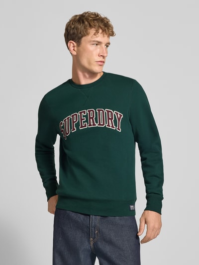 Männermodel trägt Regular Fit Sweatshirt aus Baumwoll-Mix mit Logo-Stickerei von Superdry in Dunkelgrün, Nahaufnahme Oberkörper.