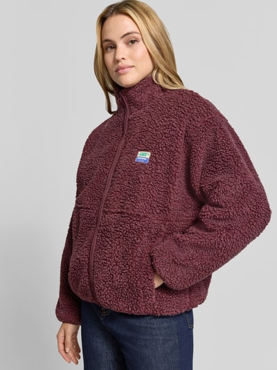 Frauenmodel trägt bordeauxrote Regular Fit Jacke aus Teddyfell Modell 'Hoktown' von American Vintage, mit Stehkragen und Logo-Patch, Nahaufnahme Oberkörper. Rückenlänge 63 cm, Schulterbreite 52 cm, Brustweite 51 cm, Ärmellänge 54 cm.