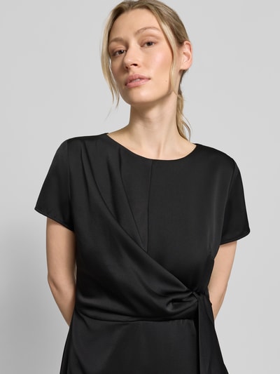 Frauenmodel trägt knielanges Cocktailkleid mit Knotendetail und Reißverschluss von Rinascimento, taillierter Schnitt, Rundhalsausschnitt, satter schwarzer Stoff mit glatter Textur, Damen, Nahaufnahme Oberkörper. Rückenlänge bei Größe S: 105 cm, Brustweite bei Größe S: 46 cm, Ärmellänge bei Größe S: 17 cm.
