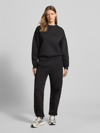 Frauenmodel trägt schwarzen Basic Sweater mit Rundhalsausschnitt und Bündchen am Ärmel von Review, Oversized, Logo-Stickerei, Ganzkörperansicht von vorne.