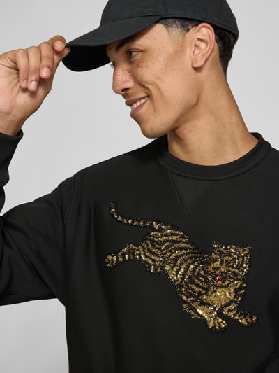 Mannenmodel draagt sweatshirt uit puur katoen van Kenzo met motiefprint van pailletten, recht pasvorm, opstaande kraag, ribboorden, ruglengte maat M 66 cm, zwart met goud en rood, detailopname bovenlichaam.