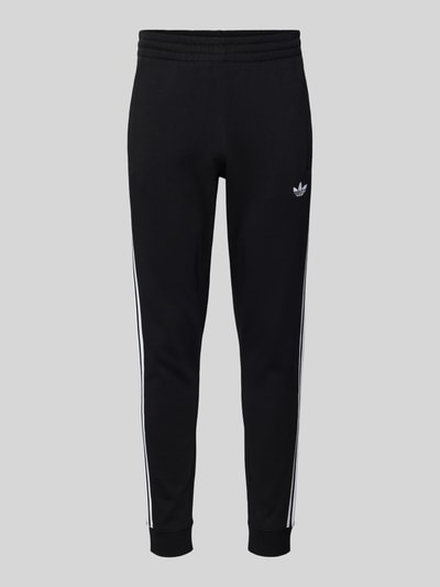Produktfoto, Sweatpants mit elastischem Bund von adidas Originals aus schwarzem Material-Mix mit Stretch-Anteil, Label-Stitching, französische Eingrifftaschen, lockerer Schnitt, Innenbeinlänge 75 cm bei Größe M, weiße Streifendetails.