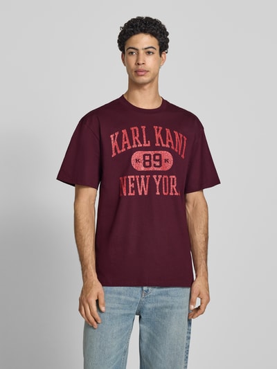 Männermodel trägt Oversized Fit T-Shirt mit Logo-Print Modell 'Heritage' von KARL KANI aus reiner Baumwolle, Rundhalsausschnitt, Bordeaux, Ganzkörperansicht von vorne.