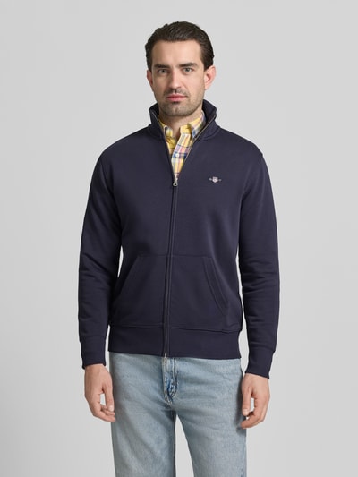 Męski model nosi Regular Fit Sweatjacke mit Stehkragen Modell 'Gield' od Gant, granatowa bluza z bawełnianego miksu, logo, zamek, kieszeń kangurka, elastyczne wykończenia, długość tyłu 69 cm, długość rękawa 64 cm, widok całej sylwetki z przodu.
