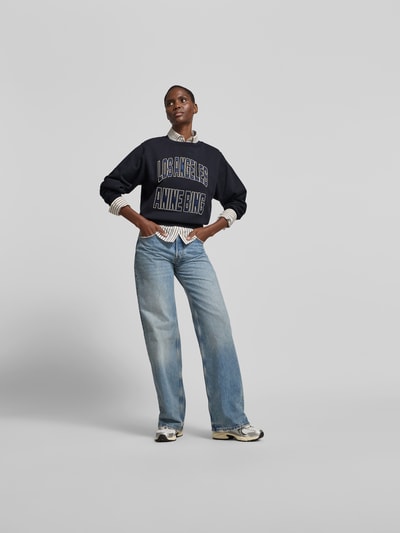 Frauenmodel trägt Sweatshirt aus reiner Baumwolle von Anine Bing in Schwarz mit Label-Detail, lockere Passform, Rundhalsausschnitt, elastische Abschlüsse, kombiniert zu Jeans und Sneakern, Rückenlänge bei Größe S 58 cm, Ganzkörperansicht von vorne.