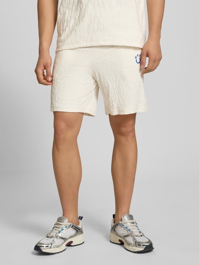 Männermodel trägt Regular Fit Shorts aus Frottee von HUGO in Creme mit Frottee-Optik, Logo-Stitching, Tunnelzug, seitlichen Eingrifftaschen, aufgesetzter Tasche hinten, Ganzkörperansicht von vorne.