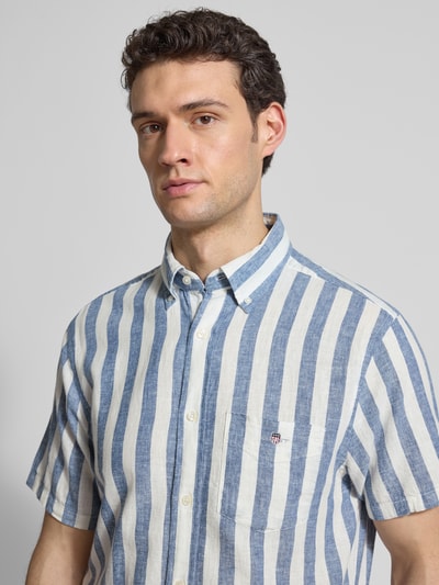 Męski model nosi Regular Fit Freizeithemd aus Leinen-Baumwoll-Mix von Gant, niebiesko-białe paski, kieszeń na piersi, krój regular fit, button-down kołnierzyk, delikatna tekstura tkaniny, długość tyłu w rozmiarze M: 77 cm, szerokość ramion w rozmiarze M: 42 cm, długość rękawa w rozmiarze M: 24 cm, zbliżenie na górną część ciała z przodu.