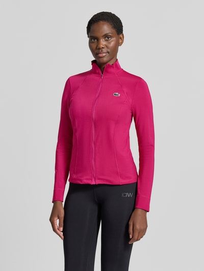Frauenmodel trägt Slim fit Trainingsjacke mit Logo-Patch von Lacoste in Pink, glatte Struktur, schwarze Hose, Ganzkörperansicht von vorne, Damen Trainingsjacke von Lacoste Sport.