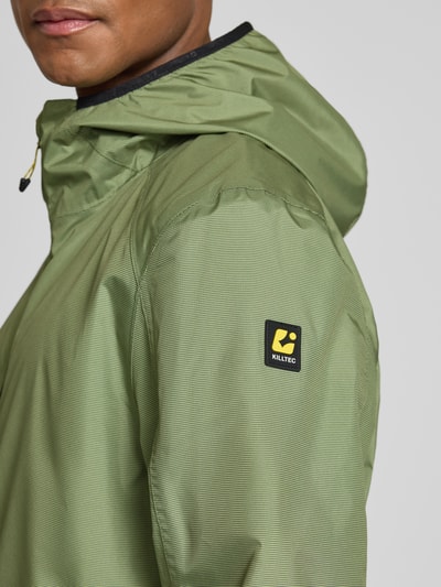 Männermodel trägt Killtec Jacke mit Kapuze Modell 'KOS' in Grün, Label-Patch, feine Struktur des Materials, Nahaufnahme Oberkörper. Rückenlänge 74 cm, Schulterbreite 52 cm, Brustweite 58 cm, Ärmellänge 68 cm, Gerader-Schnitt, Seitliche Reißverschlusstaschen, Elastische Ärmelabschlüsse.