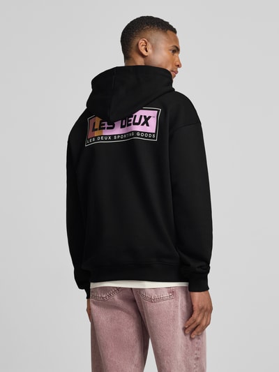 Męski model nosi Hoodie mit Logo-Print Modell 'Chase' od Les Deux z czarnej bawełny, luźny krój, nadruk z tyłu, perspektywa całej sylwetki z tyłu.