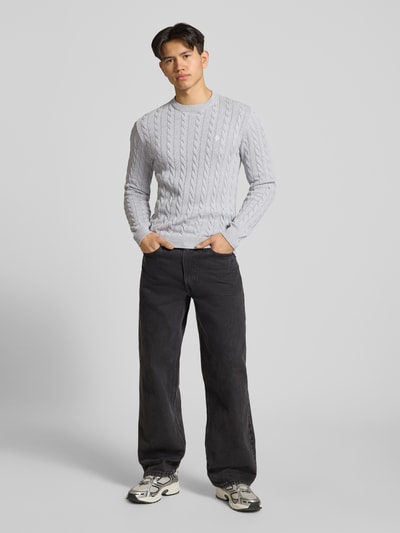 Męski model nosi Strickpullover mit Zopfmuster und Logo-Stitching od REVIEW, bawełniana mieszanka, krój modern fit, szarość, splot warkoczowy, ściągacze, przód, ujęcie całej sylwetki.