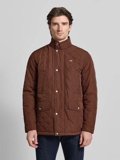 Męski model nosi Regular Fit Steppjacke mit Logo-Stitching Modell 'Windcheater' od Gant, brązowa kurtka pikowana ze stójką, wpuszczane i kieszenie z patką, z efektem pikowania, wyhaftowane logo, długość tyłu 76 cm, szerokość ramion 49 cm, szerokość klatki piersiowej 56 cm, długość rękawów 66 cm, widok na całe ciało z przodu.