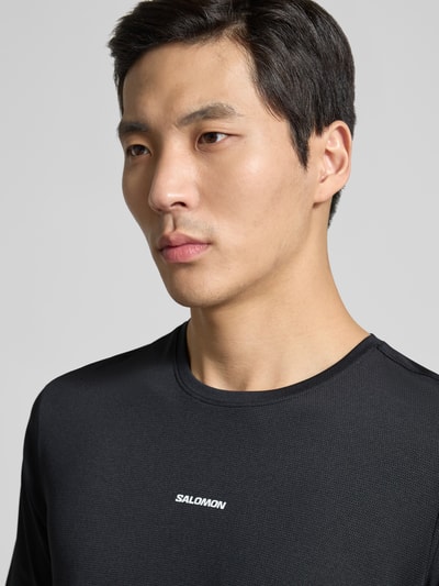 Männermodel trägt T-Shirt mit Rundhalsausschnitt und Logo von Salomon, Regular-Fit, schwarze Struktur, Nahaufnahme Oberkörper, Rückenlänge 69 cm bei Größe M.