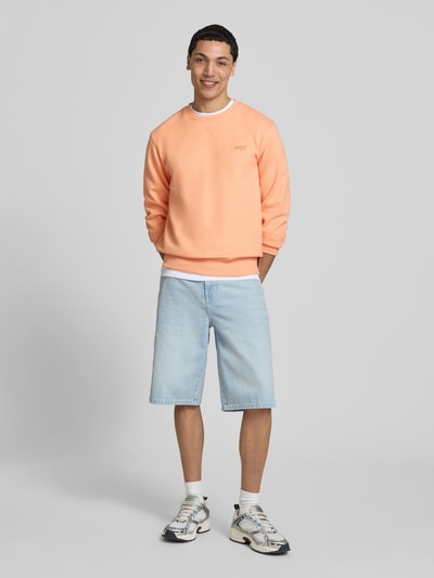Männermodel trägt Sweatshirt mit Rundhalsausschnitt und Label-Stitching von Superdry in Pfirsich, Baumwoll-Mix, Regular-Fit, Ganzkörperansicht von vorne.