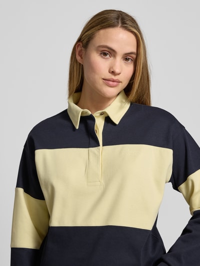 Modelka kobieca nosi Sweatshirt mit Polokragen und Label-Stitching od Gant, żółto-granatowe pasy, bawełna, relaxed fit, zbliżenie na górę ciała.