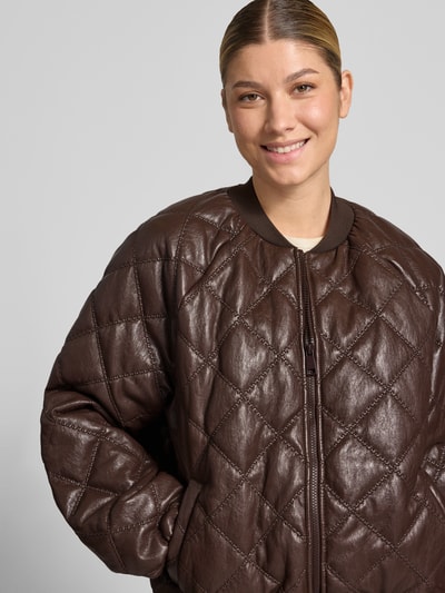 Modelka nosi Regular Fit Jacke in Stepp-Optik od Rich & Royal z materiału skóropodobnego, krój regular fit, z efektem pikowania, stójka, zamek błyskawiczny, kolor brązowy, Szerokość ramion w rozmiarze S: 43 cm, Szerokość klatki piersiowej w rozmiarze S: 52 cm, Długość tyłu w rozmiarze S: 61 cm, Długość rękawów w rozmiarze S: 46 cm, zbliżenie na górną część ciała modelki.