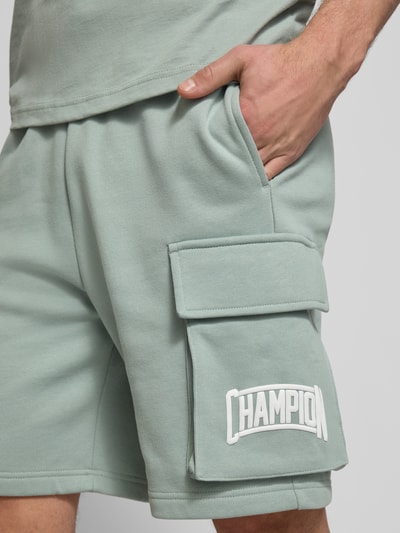 Männermodel trägt Sweatshorts mit Cargotaschen und Logo von CHAMPION in Hellgrün, aus Baumwoll-Mix, Nahaufnahme Oberschenkel.